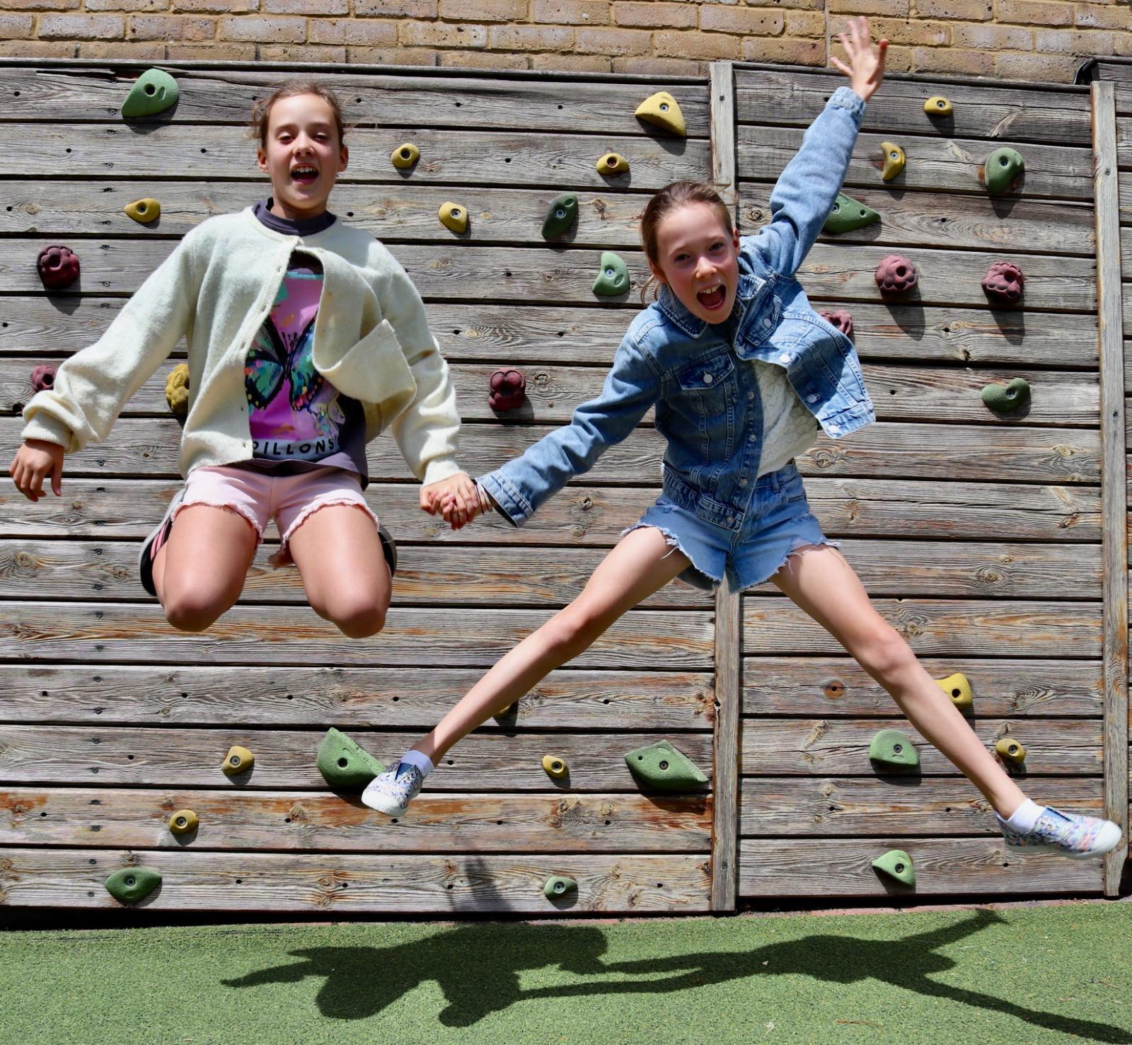 Two girls leaping in front of a climbing wall